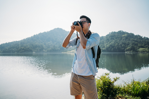 Shot of a young male traveler photographer with casual wear looking at lake scenery with camera and taking beautiful spectacular photos during  summer day.
