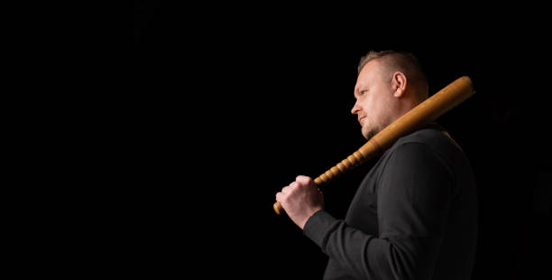 istock Young man with a baseball bat on a dark background. Bouncer, security guard, bodyguard concept. Place for text. A large man in a T-shirt stands sideways. 1369720132