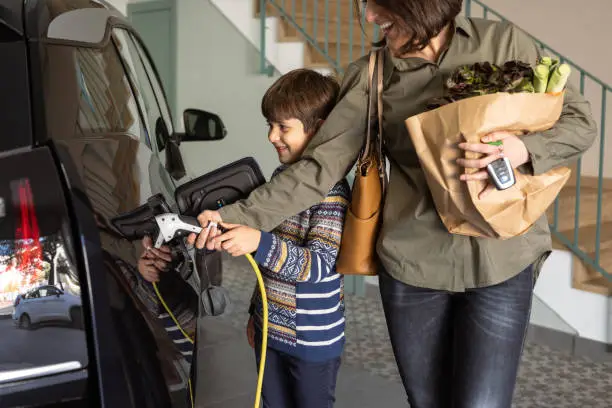 Mother and son charging electric car at home. Boy helps woman with groceries, to plug a charger yellow cable from the charging home station. Sustainable Alternative Lifestyle. Horizontal.