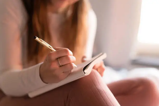 Woman is at home by herself in her apartment in the city. She is writing in her journal diary, a great way to contemplate life and mental wellbeing.