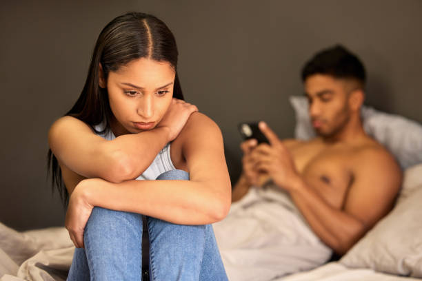 Shot of a young woman sitting on her bed and feeling upset while her boyfriend uses his cellphone behind her Is it me? human relationship stock pictures, royalty-free photos & images