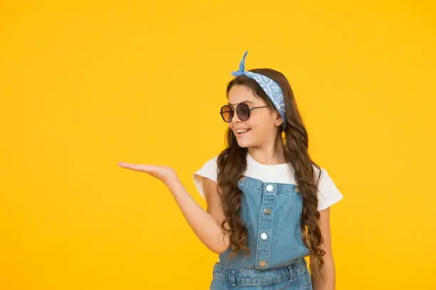 retro girl presenting product. small kid wear summer outfit. cheerful child has vintage look. headkerchief and sunglasses - summer accessory. beauty and fashion. happy childhood. copy space.