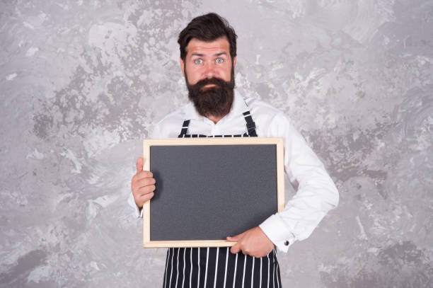 istock chalkboard with copy space for text. professional baker in apron. surprised barista with beard. advertisement and food. cook show empty board. welcome to our cafe. best chef menu. Calorie and diet 1369724020