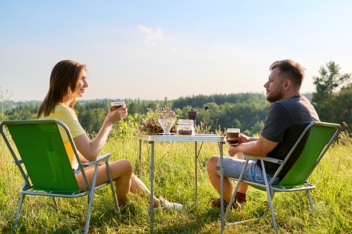 Happy middle aged couple relaxing together outdoor sitting on folding chairs at camping table. Man and woman talking, drinking fresh coffee, enjoying summer nature of wild meadow. People 40s, leisure