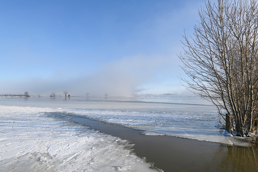 Mist rising up from the river IJssel during a cold winter morning  in the Netherlands. The water level is high and there is snow and ice on the levees on the shore of the river during this beautiful morning sunrise.