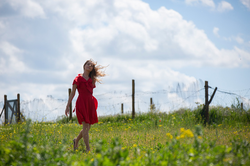 Beautiful woman in red dress walking through the flower meadow, surrounded by  old fencing mesh