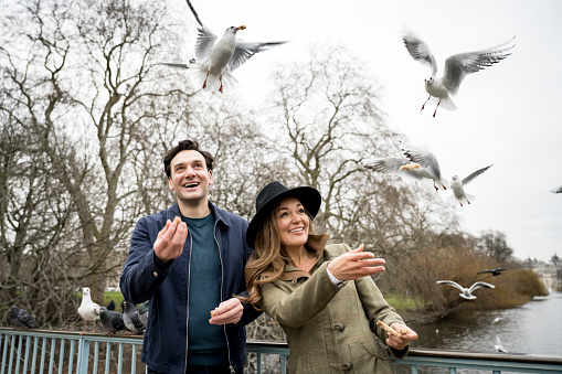 Waist-up front view of Caucasian man and woman in weekend attire standing on pedestrian bridge feeding interested group of seagulls and pigeons.