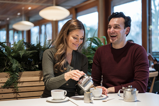 Waist-up front view of Caucasian man and woman in casual clothing smiling and laughing as they pour hot beverages and enjoy a weekend break.