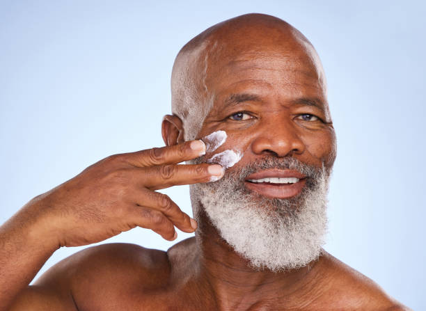 retrato de estudio de un hombre maduro que se aplica crema hidratante en la cara sobre un fondo azul - body studio shot looking at camera traditional culture fotografías e imágenes de stock