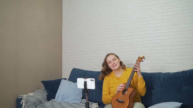 Woman conducts an online lesson and teaches students to play the ukulele. Holds guitar in hands and tells people about it via live video link. Blogging, isolation, online training