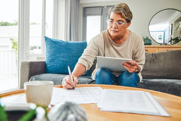 scatto di una donna matura che usa un tablet digitale mentre passa attraverso le pratiche burocratiche a casa - pension foto e immagini stock