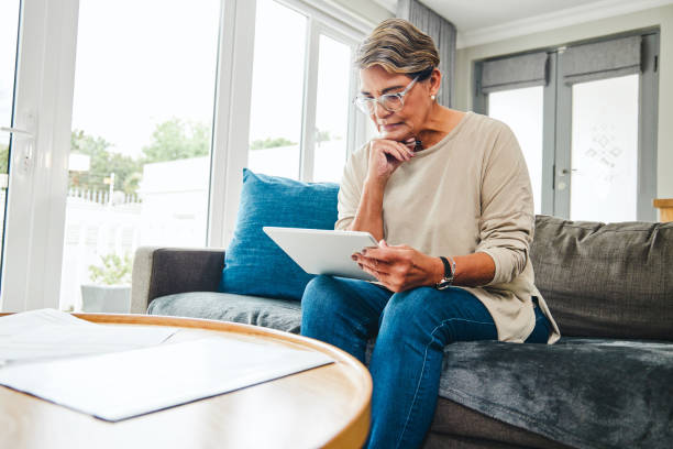 Shot of a mature woman using a digital tablet while going through paperwork at home