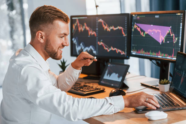 istock Using laptop. Young businessman in formal clothes is in office with multiple screens 1369747902