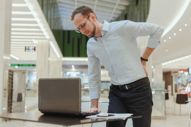 Man holds on to his back, suffers from lower back pain, standing near a desktop with a computer. Sedentary work office worker