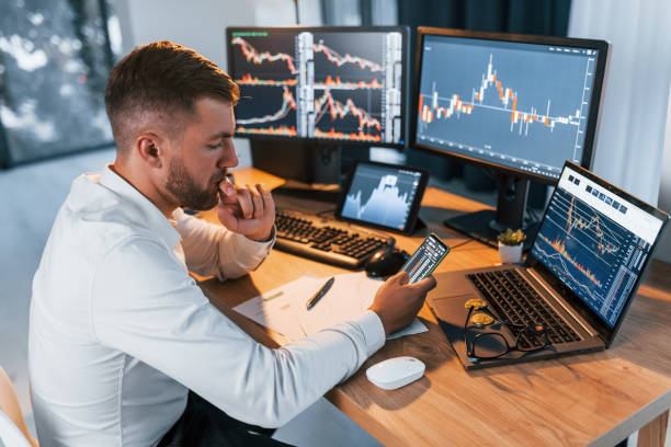 istock Side view. Young businessman in formal clothes is in office with multiple screens 1369749742