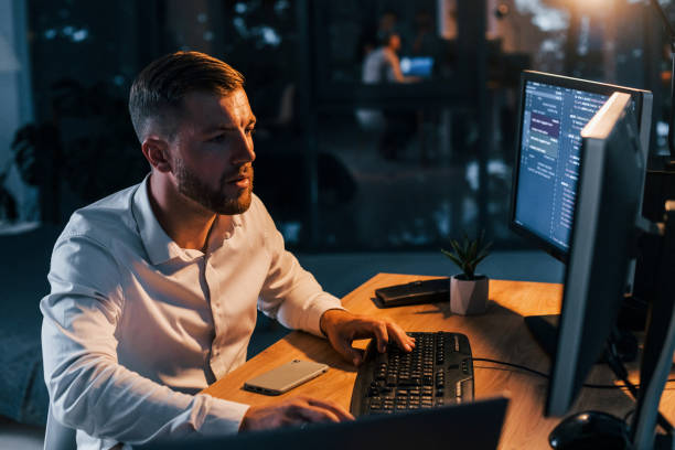 istock Working at evening time. Young businessman in formal clothes is in office with multiple screens 1369750543