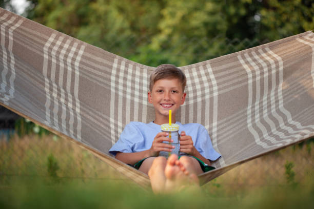 Cute smiling caucasian boy drinking water and having fun on hammock in backyard or outdoor. Summer active leisure for children. Kid swinging and relaxing in hammock.