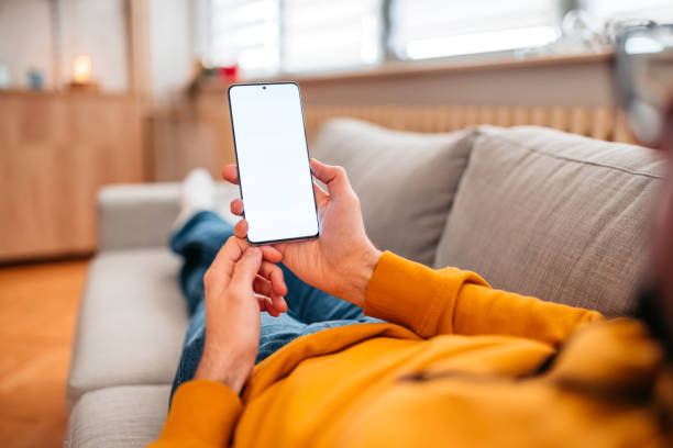 Young man using mobile phone at home Young man using mobile phone at home. Lying on the sofa and enjoying the free time. man looking at phone over shoulder stock pictures, royalty-free photos & images
