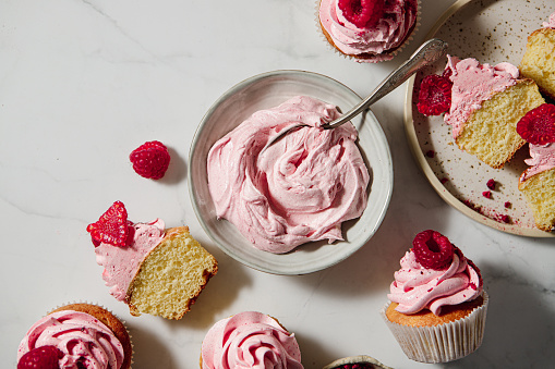 Top view of a pink whipped cream in a bowl with half sliced raspberry cupcakes on table. Homemade raspberry cupcakes on kitchen table.