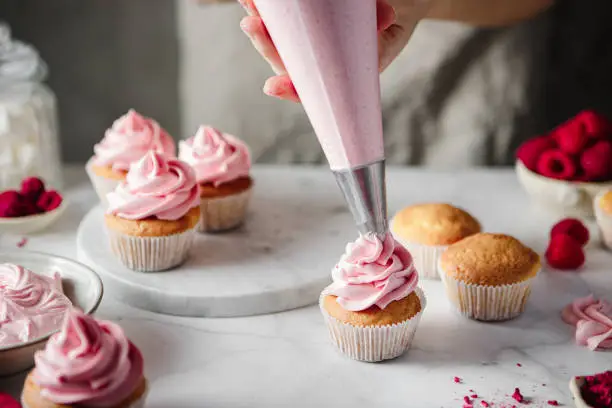 Close-up of a woman decorating muffins with pink whipped creamon the kitchen counter.  Female hands making raspberry cupcakes in kitchen.