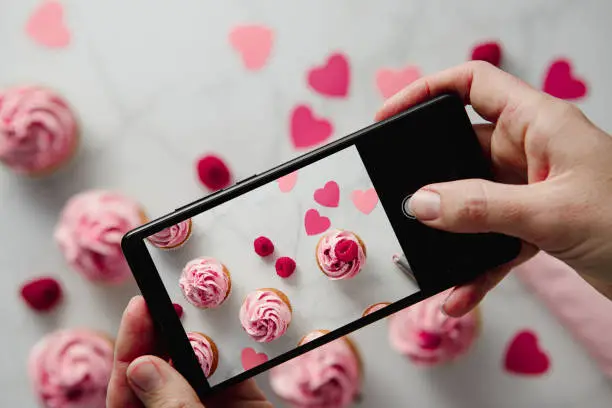 Photo of Woman photographing raspberry cupcakes with paper hearts on table