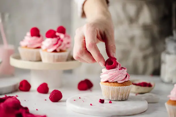 Cropped shot of a woman preparing delicious raspberry cupcakes on the kitchen counter.  Female hand decorating a pink cupcake with a raspberry topping.