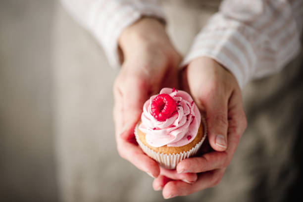 Cropped shot of a woman holding raspberry cupcake. Close-up of a female hand holding delicious pink cupcake with whipped cream and raspberry topping.
