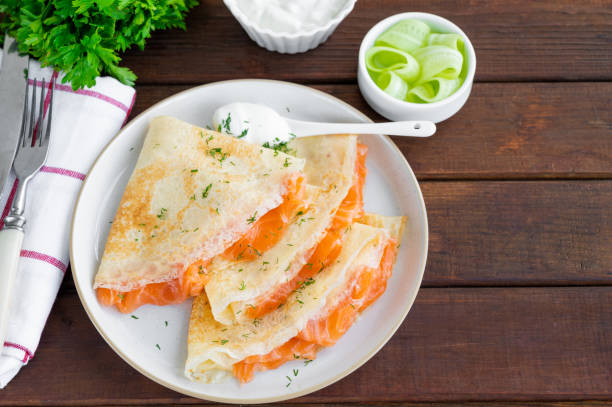 frittelle sottili o crepes con salmone affumicato, crema di formaggio, cetriolo e aneto su uno sfondo di legno scuro. messa a fuoco selettiva. - salted foto e immagini stock