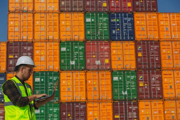 istock Engineer man with yellow crash helmet and worker west checking cargo freights in front of colorful cargo container stacks in shipping port 1369763177