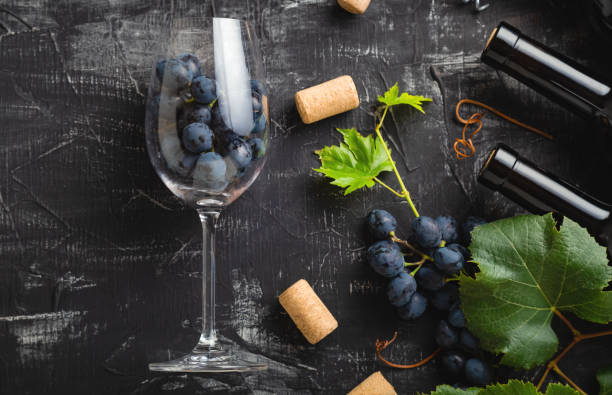 wine glass full of grapes inside. wine bottles, grape bunches with leaves and vines wine corks on dark rustic concrete background. flat lay wine composition on black stone table. long web banner. - close to food and drink yummy food imagens e fotografias de stock