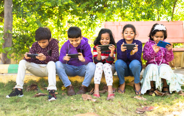 grupa dzieci zajętych korzystaniem z telefonu komórkowego podczas siedzenia w parku - koncepcja uzależnienia od smartfona, grania w gry wideo, zmniejszonej aktywności fizycznej i wpływu technologii, - video game friendship teenager togetherness zdjęcia i obrazy z banku zdjęć