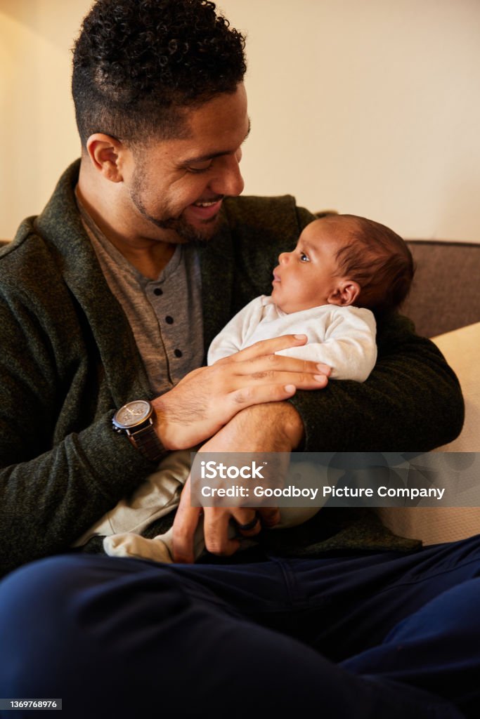 Smiling dad sitting at home and holding his baby boy in his arms Smiling young dad sitting on a sofa at home and looking at his adorable baby boy cradled in his arms Newborn Stock Photo