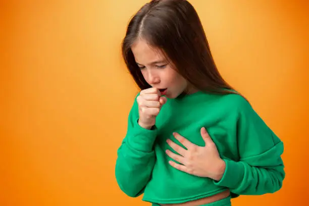 Teen girl wearing casual clothes feeling unwell and coughing against orange background in studio