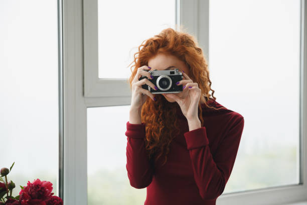 Image of young redhead lady standing near window indoors near flowers holding camera.