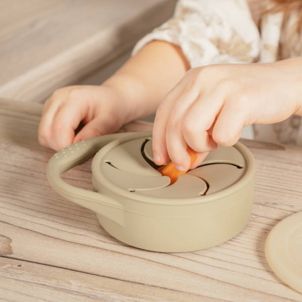 Unrecognizable child taking piece of carrot out from pastel gray silicone snack cup near lid at wooden table. Tableware. stock photo