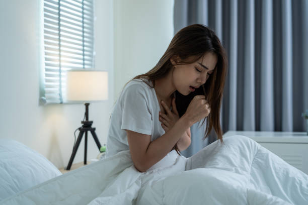 Asian beautiful girl in pajamas feel sick while sit on bed in bedroom. Attractive sleep young woman cover her mouth, feeling bad with fever and coughing after wake up on bed in the morning at home. stock photo