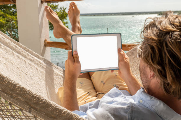 Young man relaxing in an hammock on a balcony using a digital tablet 30's man relaxing on hammock on upper floor balcony overlooking the beautiful lake of Bacalar, Mexico hammock men lying down digital tablet stock pictures, royalty-free photos & images