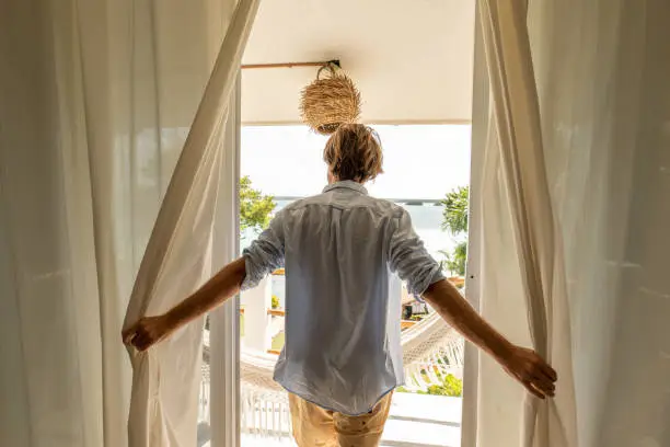 Photo of Man opening curtains in room at sunrise revealing the spectacular view, lake and garden