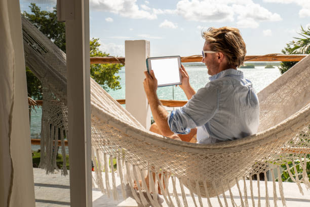 Young man relaxing in an hammock on a balcony using a digital tablet 30's man relaxing on hammock on upper floor balcony overlooking the beautiful lake of Bacalar, Mexico hammock men lying down digital tablet stock pictures, royalty-free photos & images