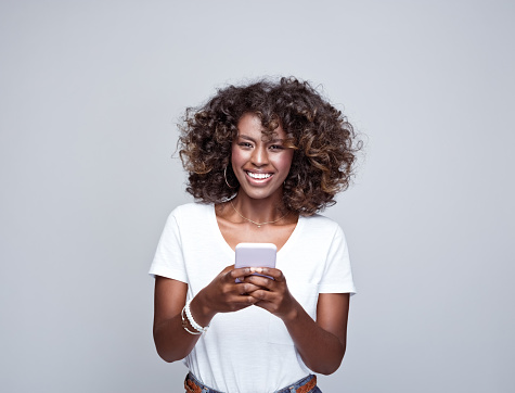 Portrait of happy, beautiful african young woman wearing white t-shirt, holding smart phone in hands and smiling at camera. Studio portrait on grey background.