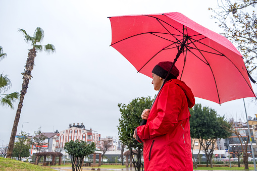 the model, who is 55 years old, has a red raincoat over a beret on her head.It's raining indoors.The season is winter.A large umbrella that plays pink in the model's hand.The model side angle is from the waist up.There are palm trees in the park