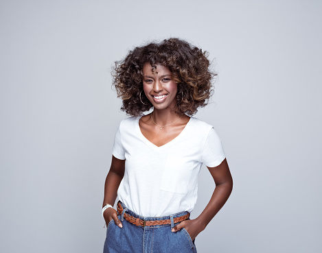 Portrait of happy, beautiful african young woman wearing white t-shirt and denim pants, standing with hands in pockets and smiling at camera. Studio portrait on grey background.