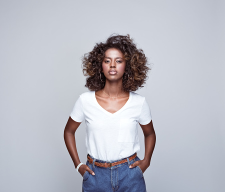 Portrait of beautiful african young woman wearing white t-shirt and denim pants, standing with hands in pockets and looking at camera. Studio portrait on grey background.