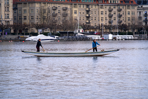 Beautiful scenic cityscape with Lake Zurich in the foreground and two women rowing on a cloudy winter afternoon. Photo taken February 3rd, 2022, Zurich, Switzerland.