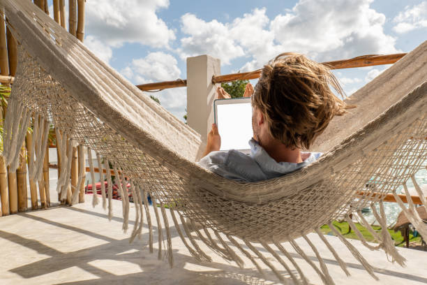 jeune homme se relaxant dans un hamac sur un balcon à l’aide d’une tablette numérique - reading beach e reader men photos et images de collection