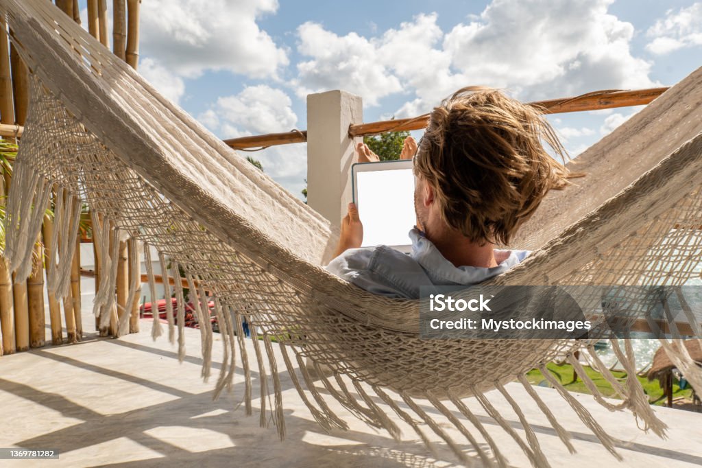 Young man relaxing in an hammock on a balcony using a digital tablet 30's man relaxing on hammock on upper floor balcony overlooking the beautiful lake of Bacalar, Mexico Awe Stock Photo