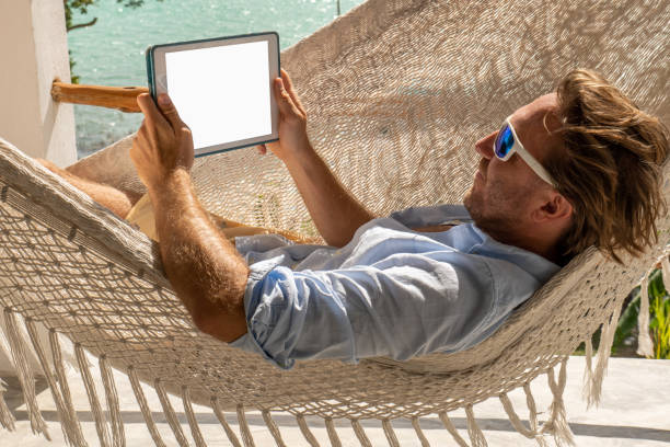 jeune homme se relaxant dans un hamac sur un balcon à l’aide d’une tablette numérique - reading beach e reader men photos et images de collection