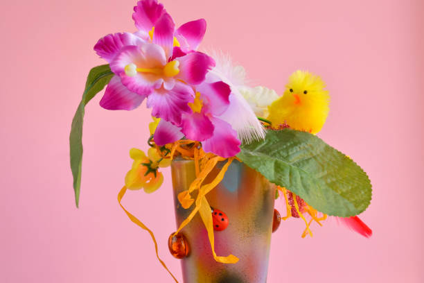 Easter composition with artificial orchid flowers and a yellow chick in a ceramic pot on a pink background, festive home decor