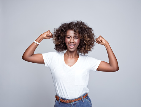 Portrait of happy, beautiful african young woman wearing white t-shirt and denim pants, standing raised arms and smiling at camera. Studio portrait on grey background.