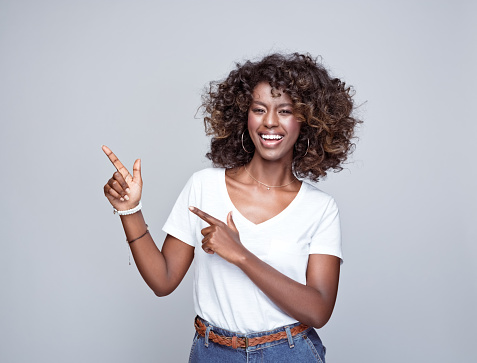 Portrait of happy, beautiful african young woman wearing white t-shirt and denim pants, pointing with index fingers at copy space and smiling at camera. Studio portrait on grey background.